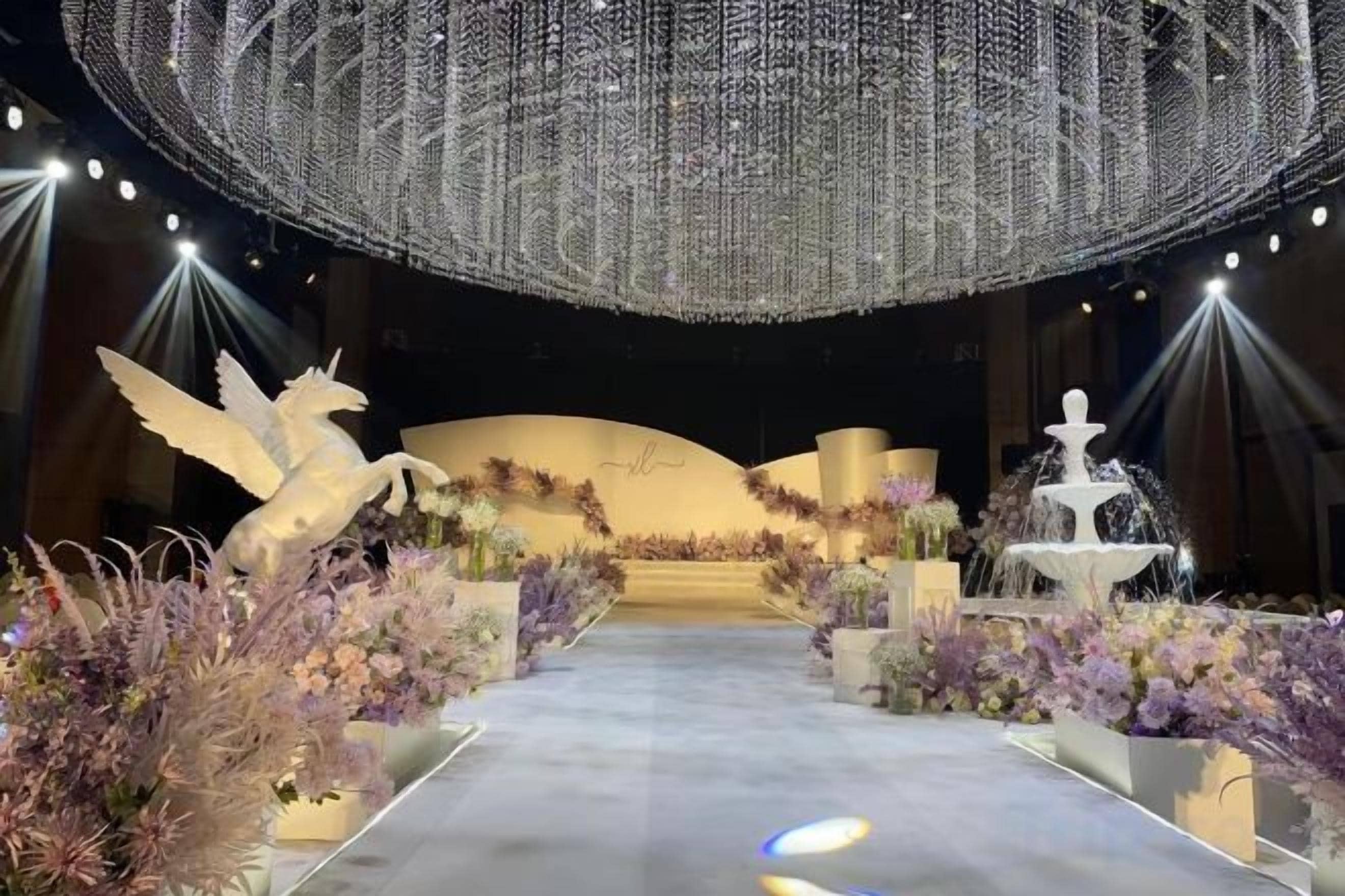 Ein festlicher Veranstaltungsraum mit einer zentralen Bühne, Blumendekorationen und einem großen Chandelier.