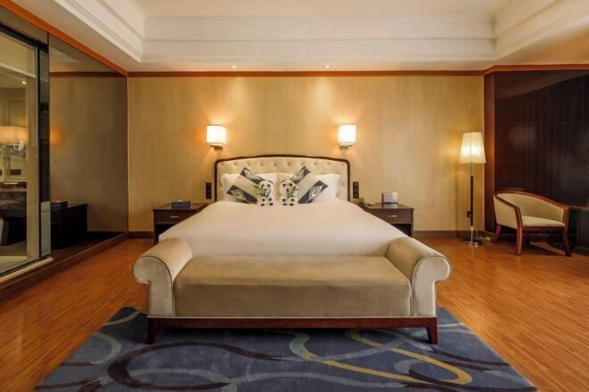 A hotel room with a large bed, two bedside lamps, a chair, and a glass partition.