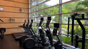 Ein Fitnessstudio mit Laufbändern, Crosstrainern und Kraftgeräten, große Fenster mit Blick auf Grünflächen.