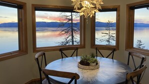 Un coin repas avec une table ronde, six chaises et un lustre. Trois grandes fenêtres offrent une vue sur un lac et des montagnes.