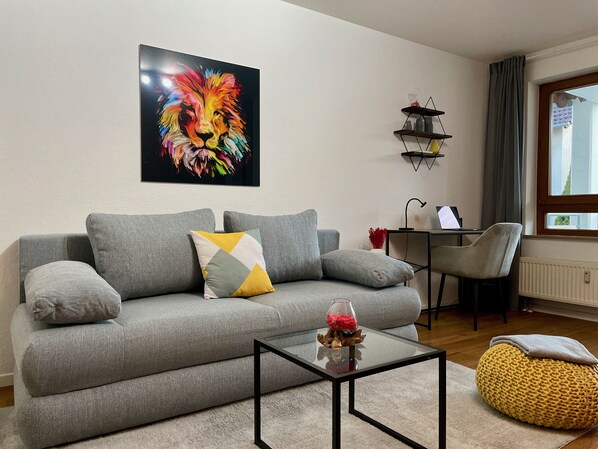A living room with a grey sofa, a glass coffee table, a colorful lion painting, a small side table, and a window with curtains.