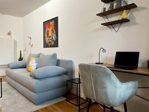 A modern living room with a grey sofa, a grey armchair, a wooden desk with a laptop, a black desk lamp, and a colorful abstract painting on the wall.