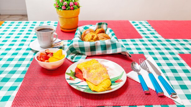 Une table recouverte d’une nappe à carreaux, un saladier rempli de fruits, une assiette avec une omelette, des croissants, une tasse de café et une plante en pot.