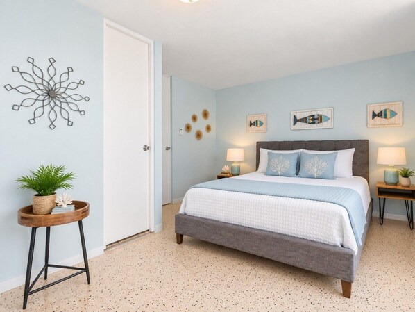 Quarto com cama, mesas de cabeceira, uma obra de arte na parede e uma pequena mesa lateral com uma planta.