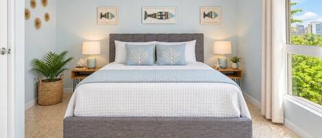 A bedroom with a bed, two bedside tables, wall art, and a view through a window.