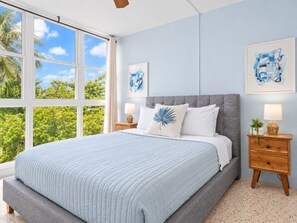 Ein Schlafzimmer mit einem großen Bett, einem Nachttisch, einem Fenster mit Blick auf Grünflächen und zwei an der Wand befestigten Kunstwerken.