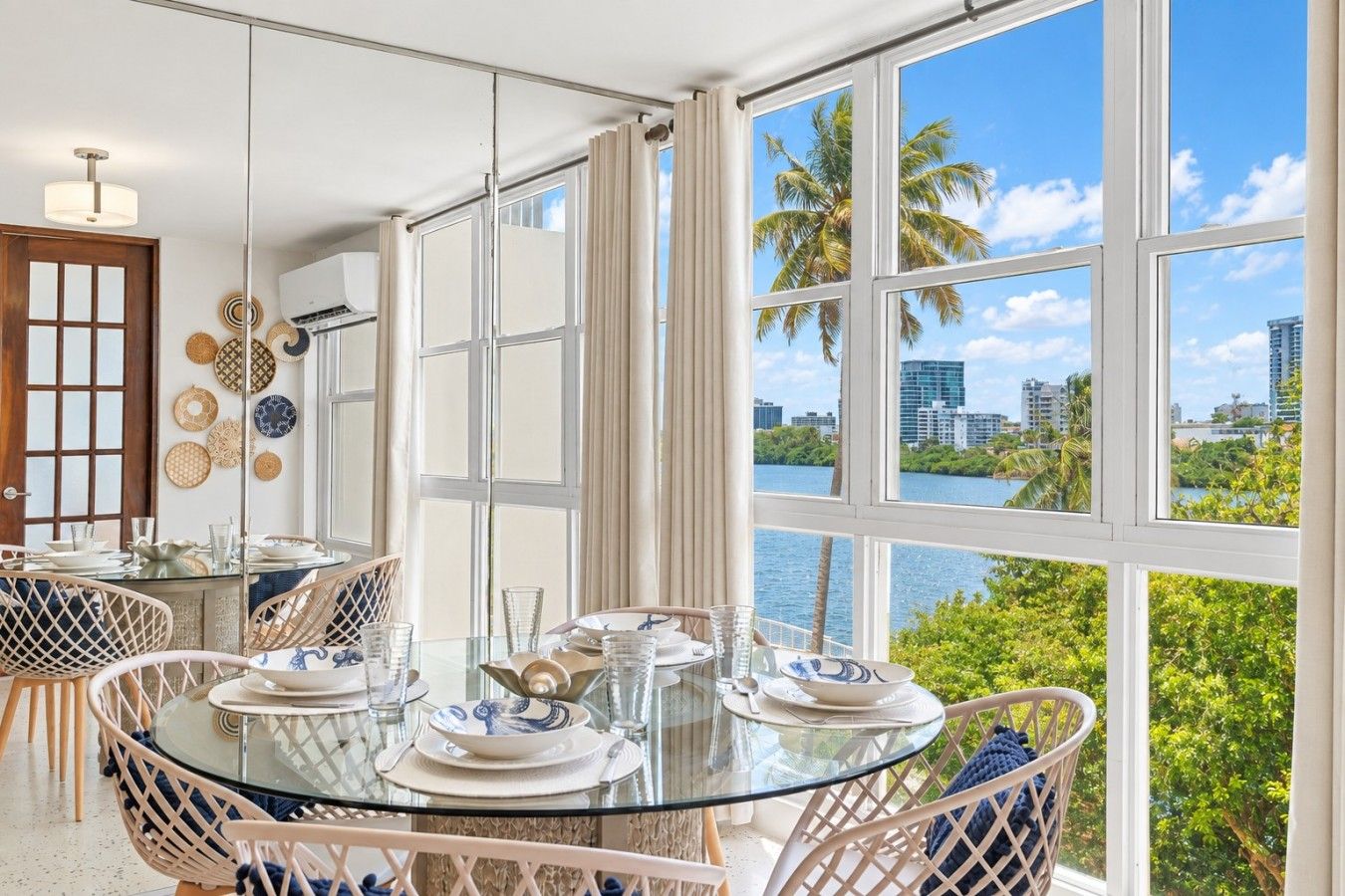 Un coin repas avec une table et des chaises en verre, une vue sur l’eau et les palmiers, et un paysage urbain en arrière-plan.