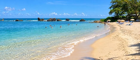 A beach with clear water, people swimming, and sun loungers under umbrellas.