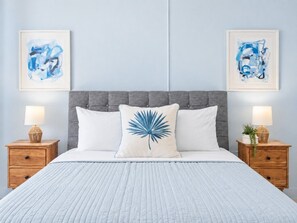 A bed with a grey upholstered headboard, two white pillows, a blue palm leaf pillow, and a white quilted bedspread. There are two wooden nightstands with lamps and a small plant on each.