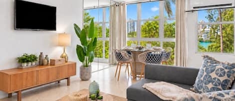 A modern living room with a sofa, a TV, a dining area with chairs, and large windows.