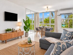 A modern living room with a sofa, a TV, a dining area with chairs, and large windows.