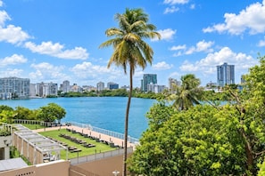 A waterfront view with a pool area, palm trees, and a city skyline in the background.