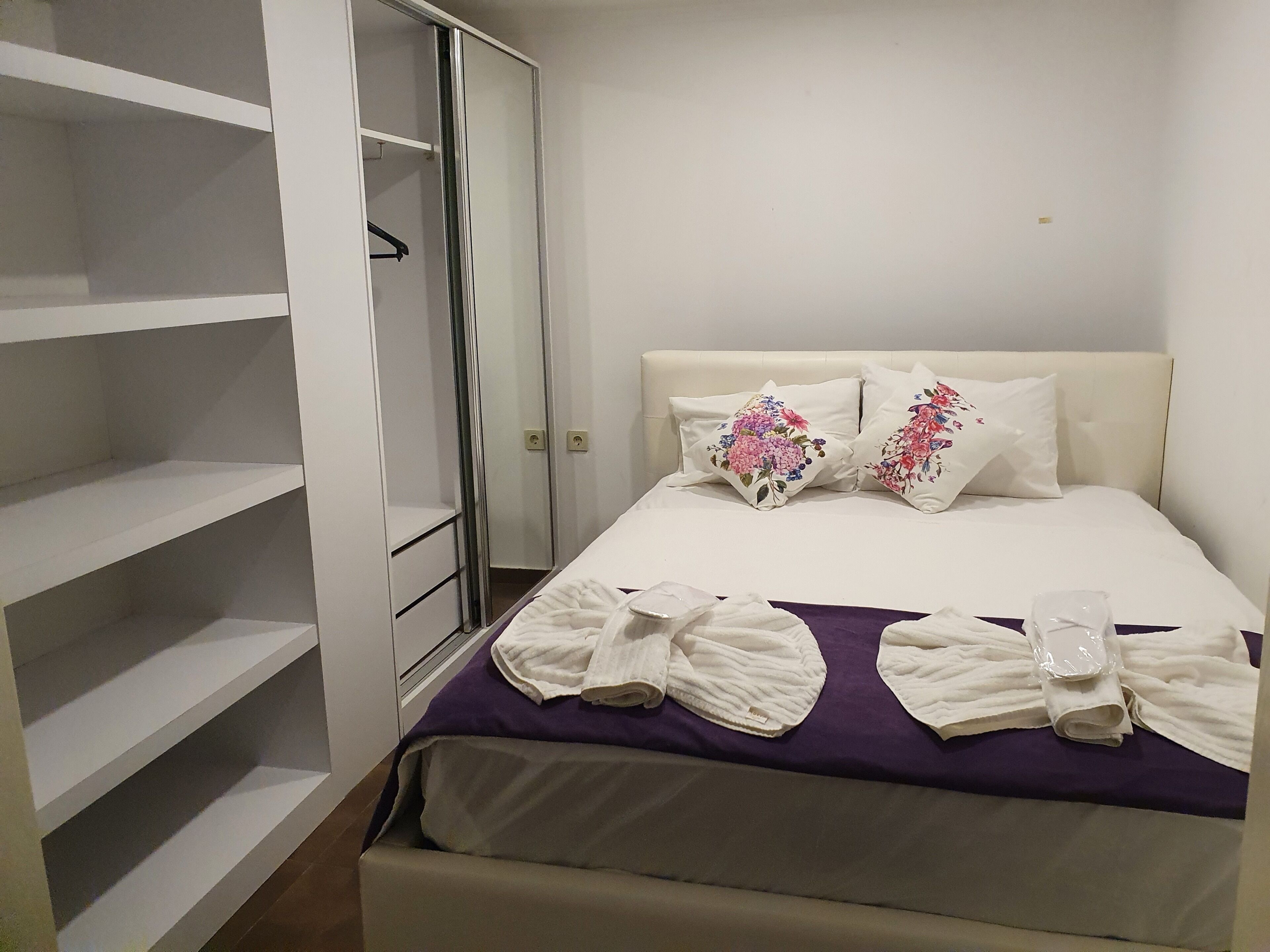 Un lit bien fait, agrémenté d’oreillers à motifs floraux, d’une couverture violette et de serviettes enroulées.