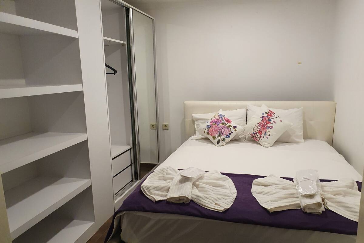A neatly made bed with floral pillows, a purple blanket, and rolled towels.