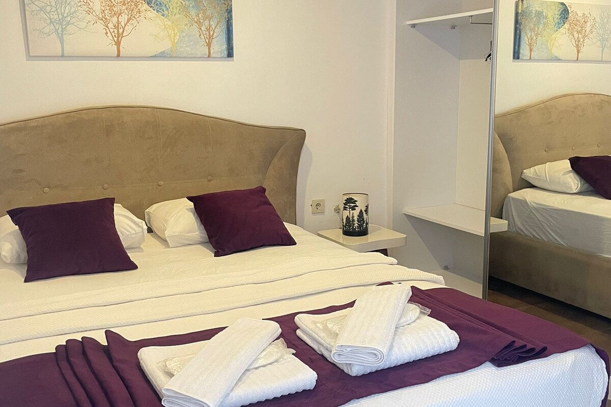 A hotel room with a large bed, a bedside table, a nightstand, a mirror, and two wall-mounted paintings.