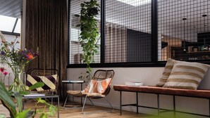 A modern living room with a wooden floor, a wicker chair, a bench, a small table, and a large window with a view of buildings.