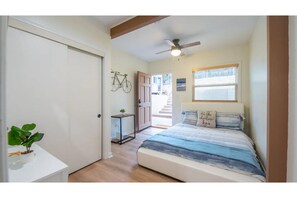Un dormitorio con cama, ventilador de techo, ventana con persianas, una puerta y una mesita con una planta.