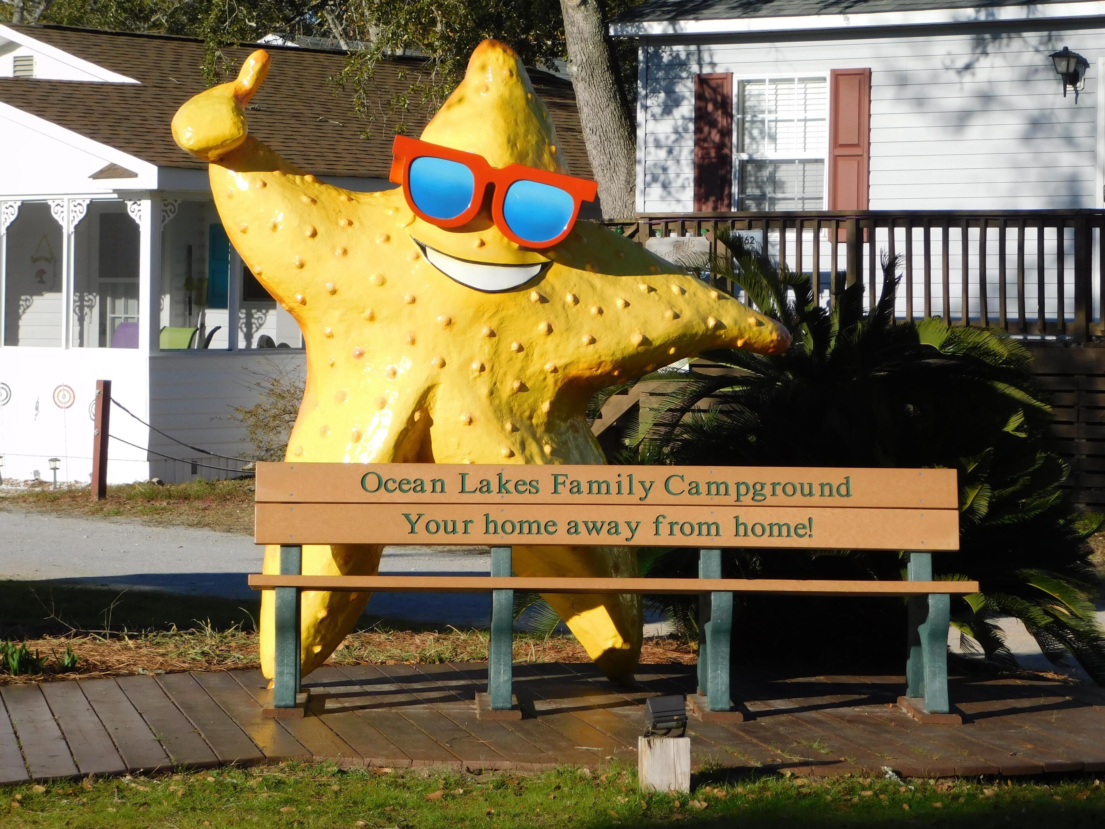 Une grande sculpture d’étoile de mer jaune portant des lunettes de soleil, un banc en bois et un panneau indiquant : « Ocean Lakes Family Campground – Votre chez-vous loin de chez vous ! »