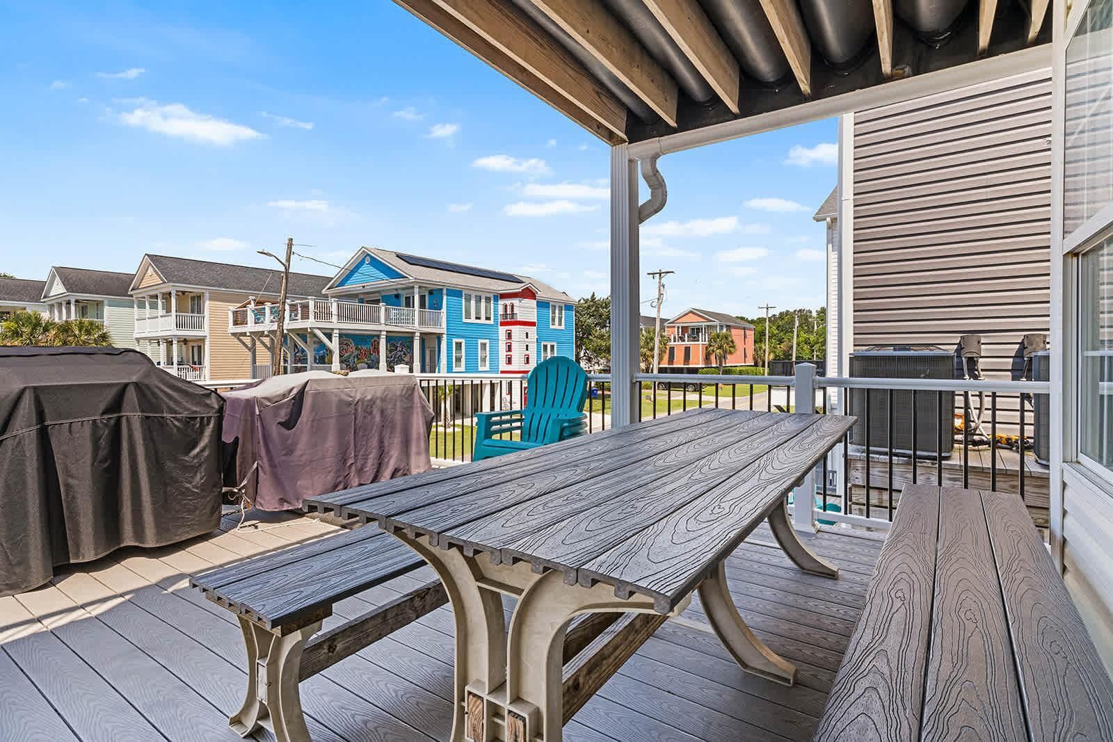 A covered outdoor area with a picnic table, barbecue grill, and a view of colorful houses in the background.