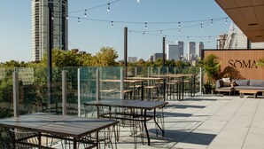 An outdoor seating area with tables and chairs, a sofa, and string lights. A tall building is visible in the background. - Brut Hotel (Tulsa)