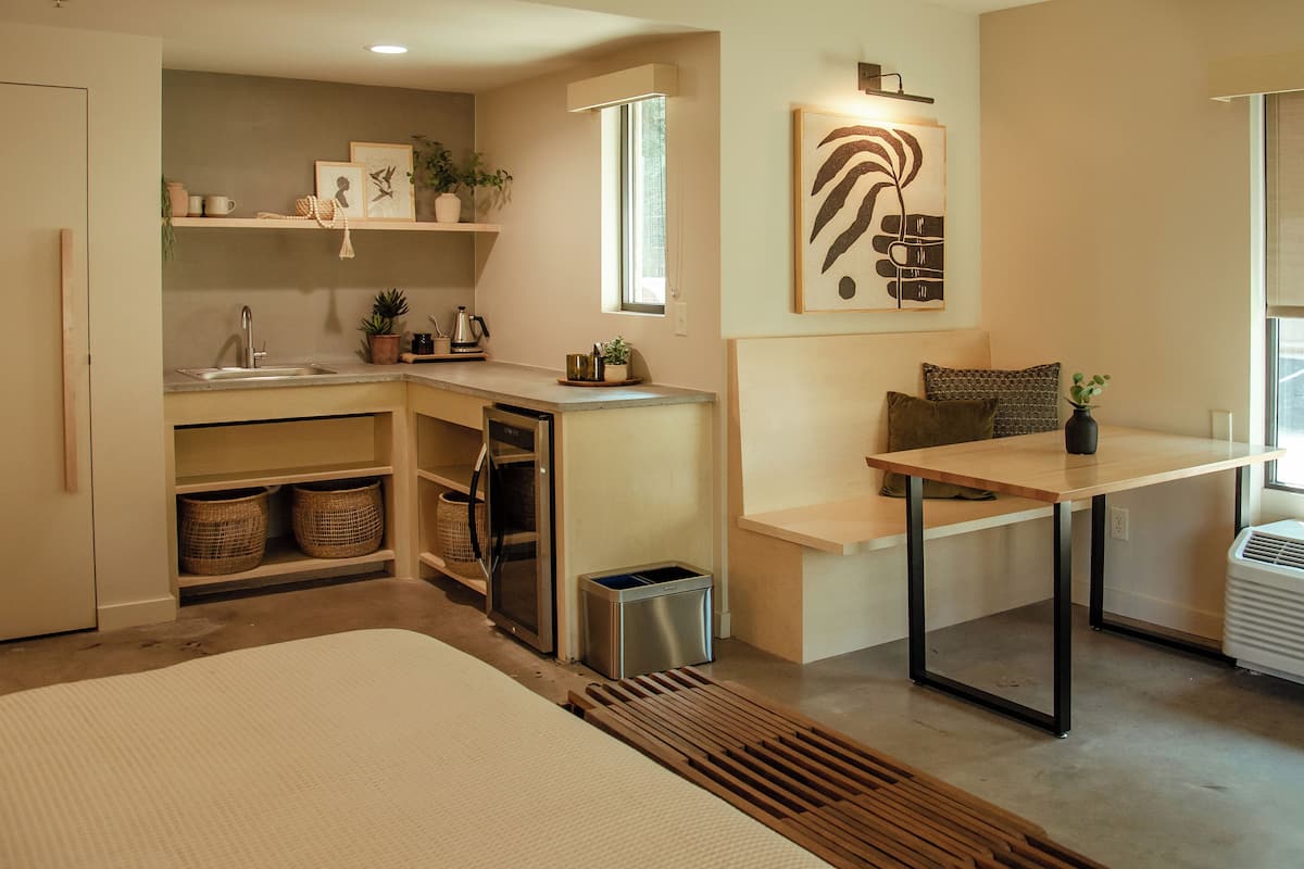 A compact kitchen with a sink, countertop, and a window. There is a small table with a vase and a pillow.