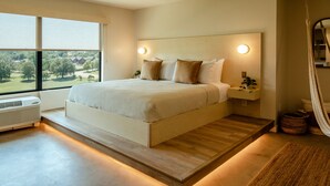 A modern bedroom with a large bed, built-in bedside tables, and a view of a green landscape through large windows. - Brut Hotel (Tulsa)