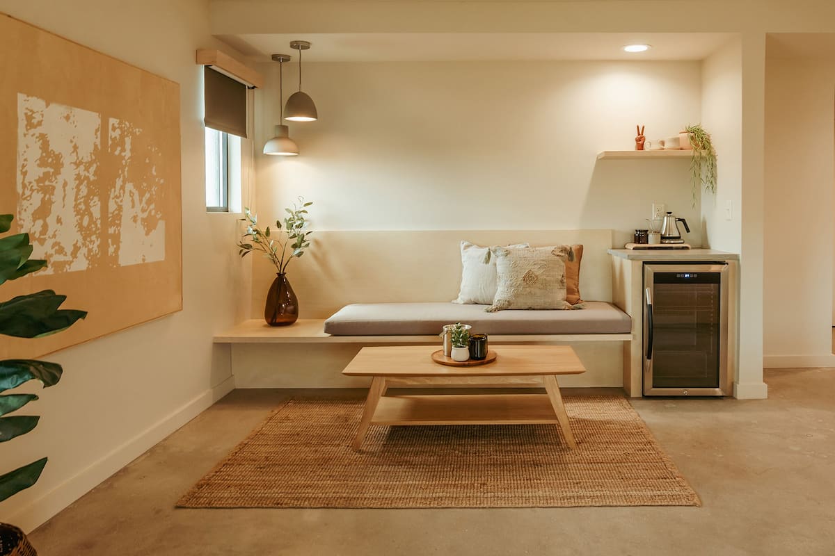 A modern living room with a sofa, a coffee table, a small kitchen area with a refrigerator, and a large plant.