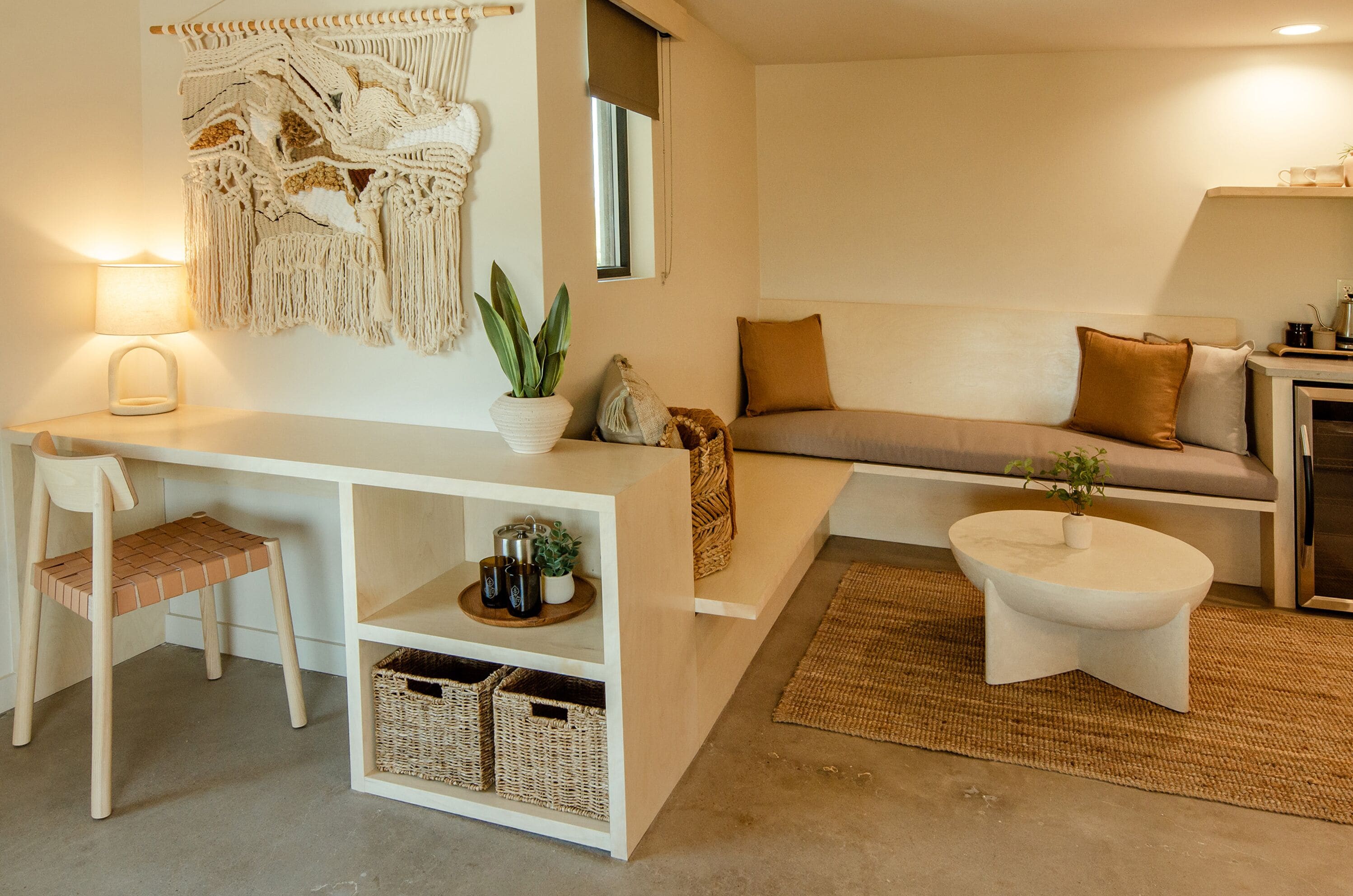A modern living room with a white desk, a cushioned bench, a round coffee table, and a macrame wall hanging.