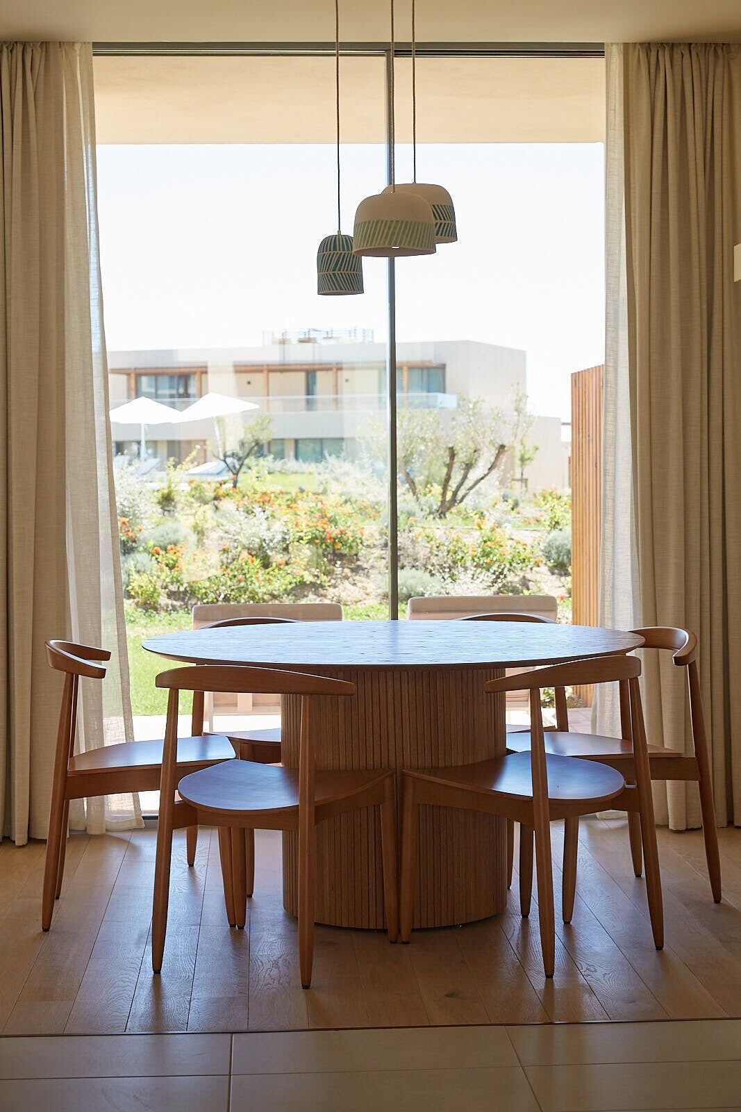 Un coin repas avec une table ronde en bois et six chaises, de grandes fenêtres avec des rideaux, et une vue sur un jardin et des bâtiments à l’extérieur.