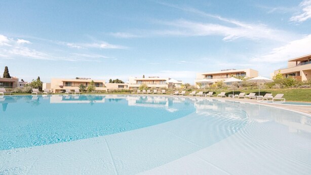 Una grande piscina con lettini e ombrelloni, circondata da edifici moderni e un cielo sereno.