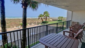 Uma varanda com vista para a praia, palmeiras e uma mesa com cadeiras.