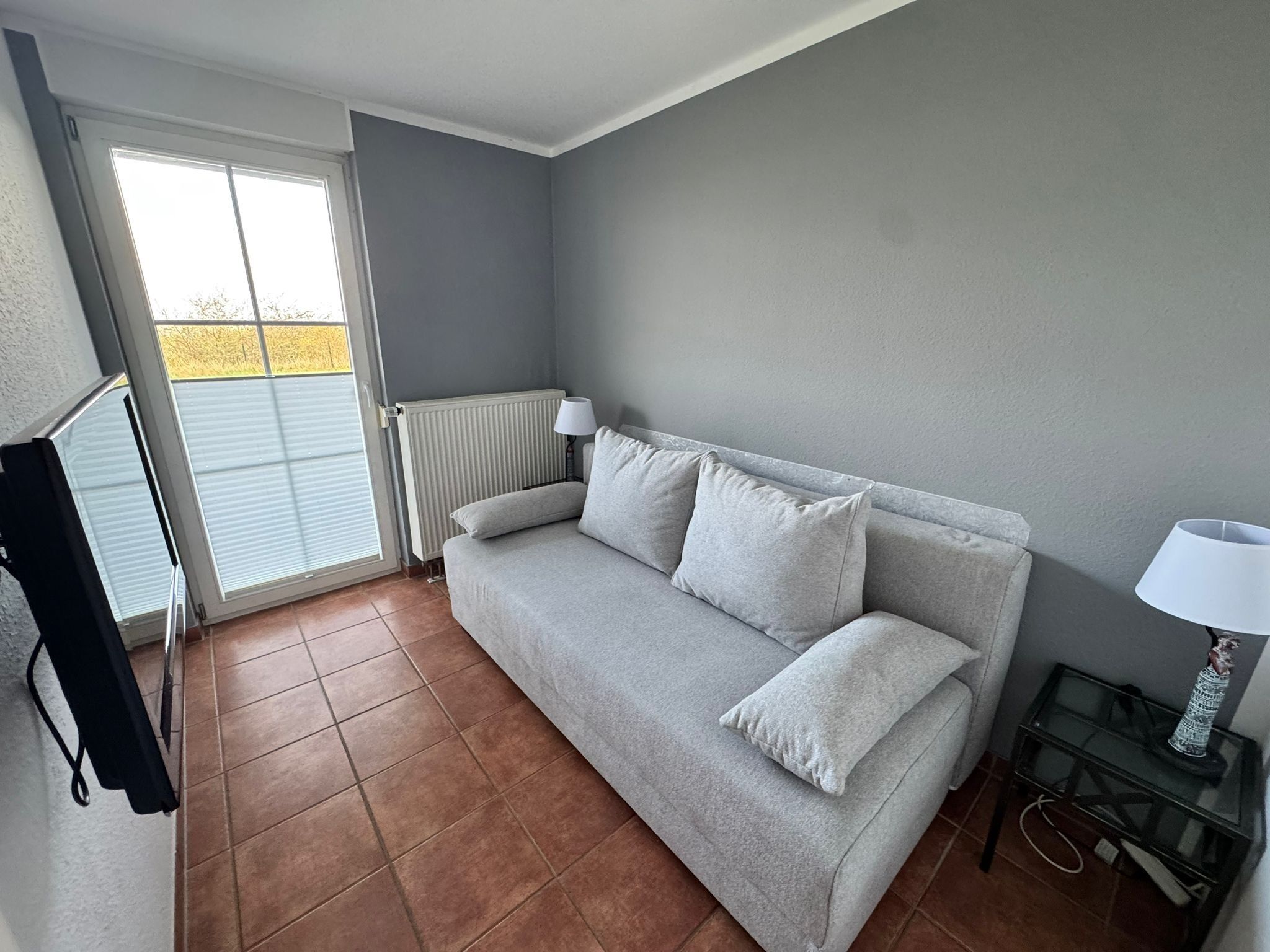 Une pièce avec un canapé gris, un radiateur, une lampe et une fenêtre avec des stores.