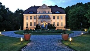 Un grand bâtiment de style classique, doté d’une façade symétrique, d’une entrée centrale et d’un jardin à la française, avec un chemin pavé menant à l’entrée.