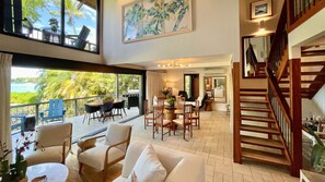 Un vaste espace de vie avec un balcon donnant sur un paysage tropical, un escalier en bois et un coin repas doté de meubles modernes.