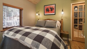 A bedroom with a checkered bedspread, a wooden headboard, two wall-mounted lamps, a framed floral artwork, and a window with blinds. - ⭐️Premier Host - Serene cabin, the best amenities, north end of James Ranch  (Durango)