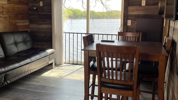 A cabin interior with a wooden ceiling, a dining table with chairs, a leather sofa, and a view of a lake through a large window.