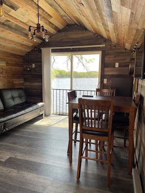 Interior de cabana com teto de madeira, mesa de jantar com cadeiras, sofá de couro e vista para um lago através de uma grande janela.
