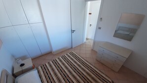 A bedroom with a bed, a bedside table, a dresser, a striped rug, and a door leading to another room. - Red House Pescoluse (Marina di Pescoluse)