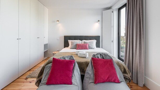 A modern bedroom with a large bed, two bedside tables, a wardrobe, and a view through a large window. - GuestReady - One Step Closer (Porto)