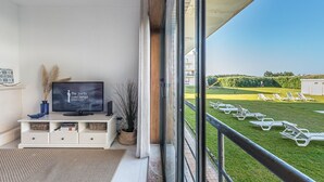 Un balcon équipé d’un téléviseur, d’une console blanche et offrant une vue sur un espace verdoyant avec des chaises longues.