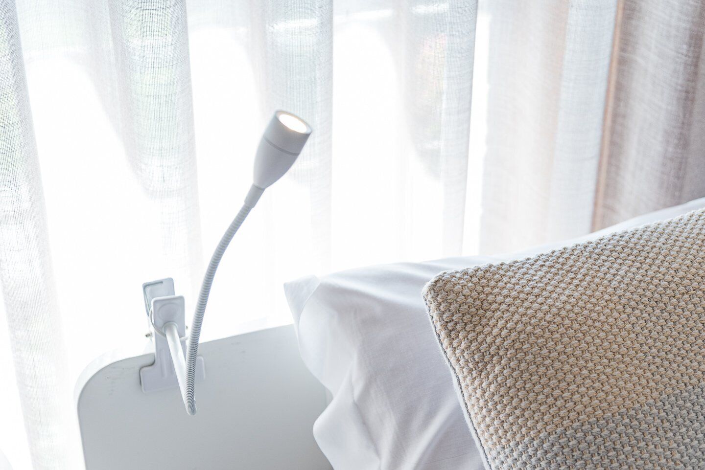 A white bedside table with a lamp, a white pillow, and a window with sheer curtains.