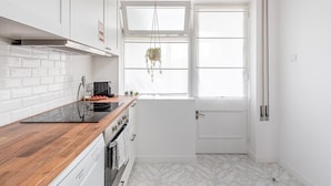 Une cuisine compacte avec des meubles blancs, un plan de travail en bois, un four encastré, une fenêtre agrémentée d’une plante suspendue et une porte dotée d’une poignée.