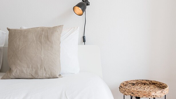 Un lit bien fait, avec du linge de lit blanc et deux oreillers, une lampe de sol noire et un tabouret en osier.