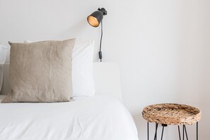 Un lit bien fait, avec du linge de lit blanc et deux oreillers, une lampe de sol noire et un tabouret en osier.