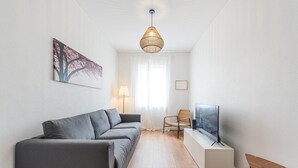 Un salon moderne avec un canapé gris, une table basse en bois, un téléviseur, un fauteuil en osier et un luminaire suspendu.