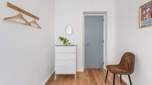 Une pièce avec un plancher en bois, une commode blanche à tiroirs, un miroir, une plante suspendue et un fauteuil marron.