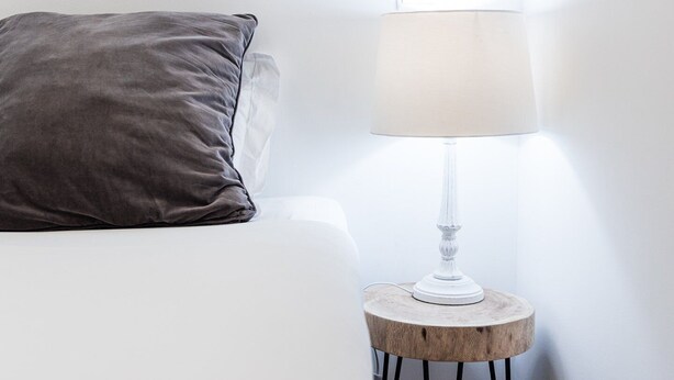 Un lit blanc avec un oreiller gris et une lampe de table transparente posée sur un tabouret en bois.