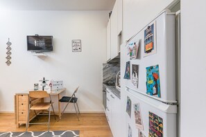Uma cozinha compacta com geladeira decorada com ímãs de viagens, uma escrivaninha de madeira com cadeira e uma televisão fixada na parede.