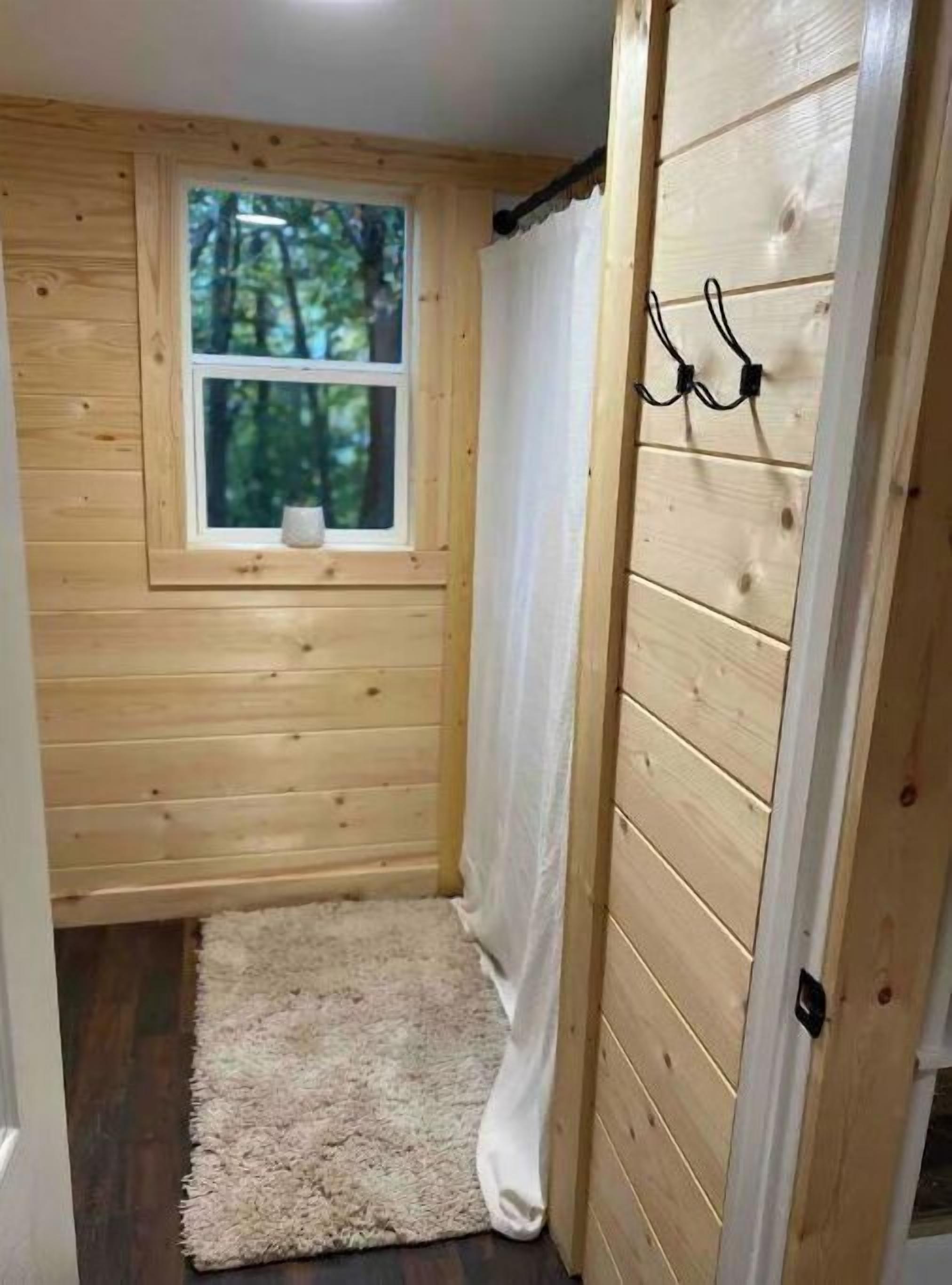 Una habitación pequeña de madera con ventana, cortina blanca, alfombra mullida y puerta con un perchero.