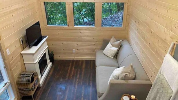 A cozy cabin interior with a sofa, a fireplace, and three windows offering a view of the forest.